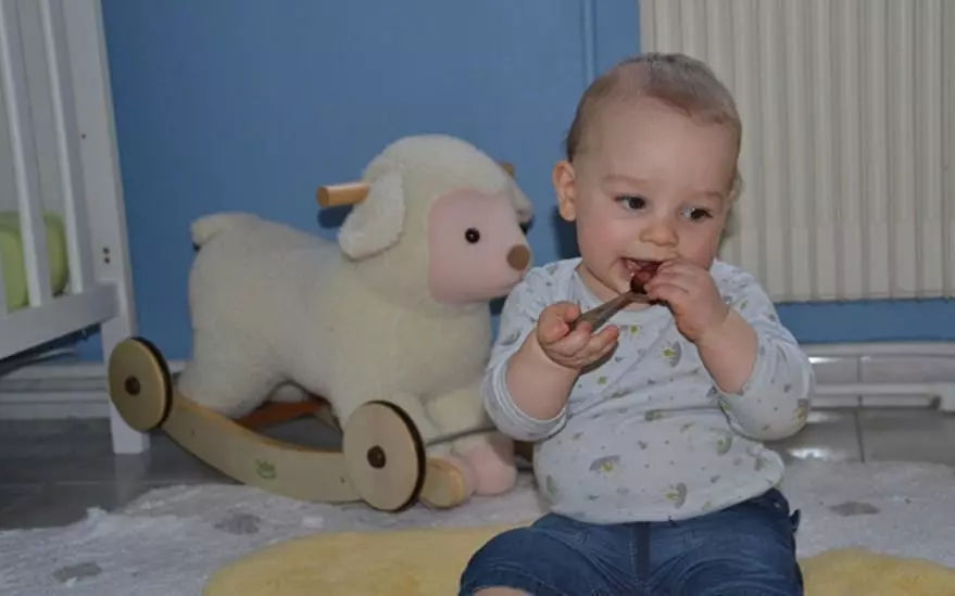 Un jeune enfant est assis sur un tapis jaune moelleux dans une chambre d'enfant, jouant pres d'un transat en forme d'agneau en peluche. La piece est dotee d'un mur bleu et d'un lit bebe blanc a l'arriere-plan, creant une atmosphere chaleureuse et adaptee aux enfants. Le transat en forme d'agneau est dote de roues en bois et d'un design doux et texture.