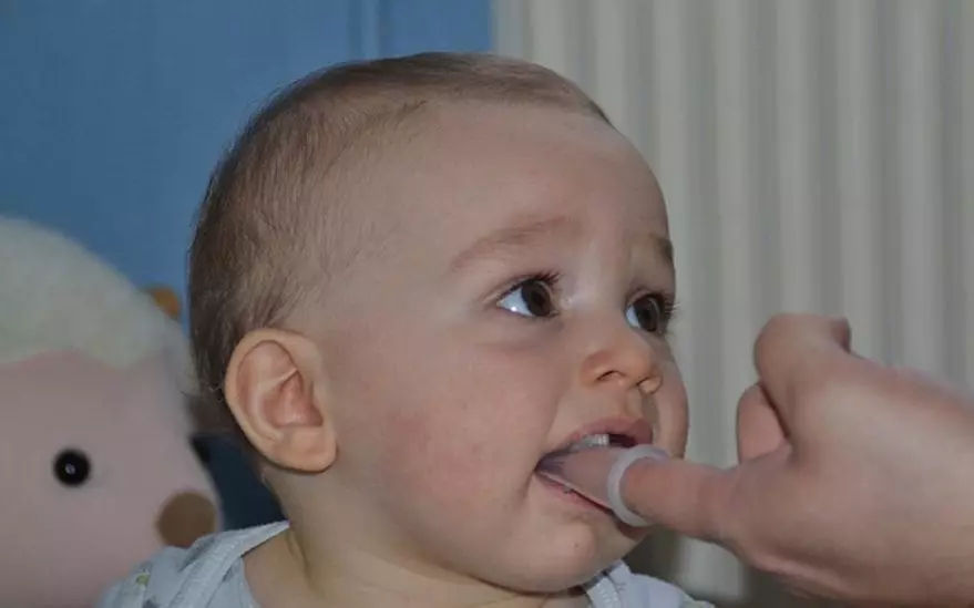 Un bebe est assis a l'interieur, avec une peluche visible en arriere-plan. Le decor semble etre une piece confortable avec un eclairage tamise et des tons neutres. Le bebe porte des vetements de couleur claire et une main est tendue pres de lui, suggerant une interaction ou un jeu.