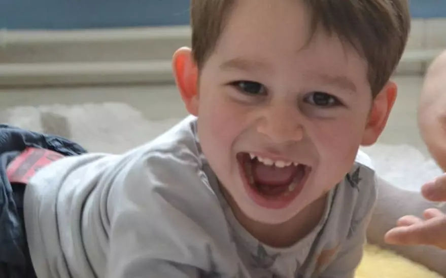 Un jeune enfant est allonge sur une surface moelleuse a l'interieur, vetu d'une chemise claire et d'un pantalon fonce. Le decor semble decontracte et familier, avec un arriere-plan flou et une main tendue a proximite. La scene degage une atmosphere detendue et ludique.