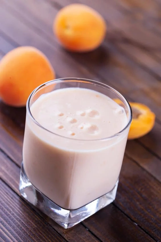 A close-up of a creamy apricot smoothie served in a square glass on a wooden table. Fresh apricots are placed nearby, adding a natural and vibrant touch to the setting. The smooth texture and light peach color evoke a refreshing and healthy beverage option.