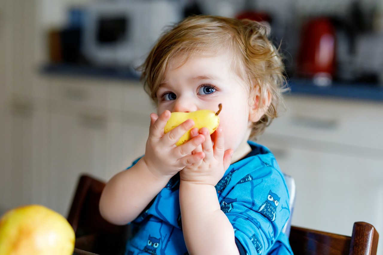 Cute adorable toddler girl eating fresh pear . Hungry happy baby child of one year holding fruit. Girl in domestic kitchen, having healthy meal snack. Smiling blond kid.