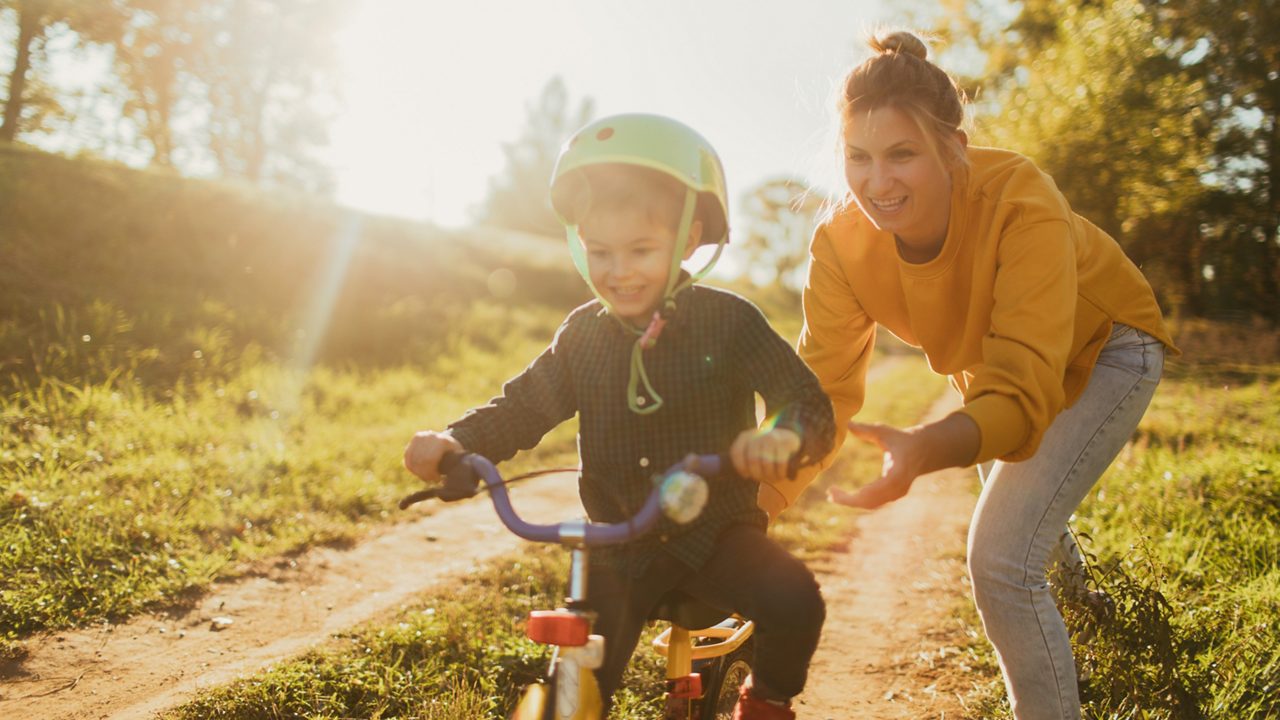 Photo of a young boy who is learning to ride a bicycle with a little help from his mother; wide photo dimensions