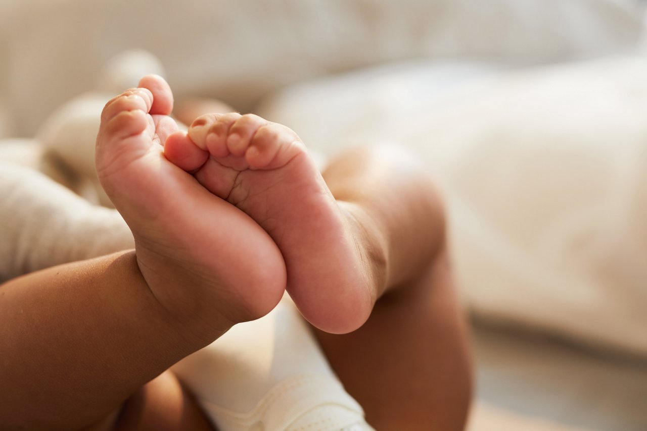 Schwangere sitzt vor ihrem Mann auf der Couch, er hat die Hände auf ihrem Bauch