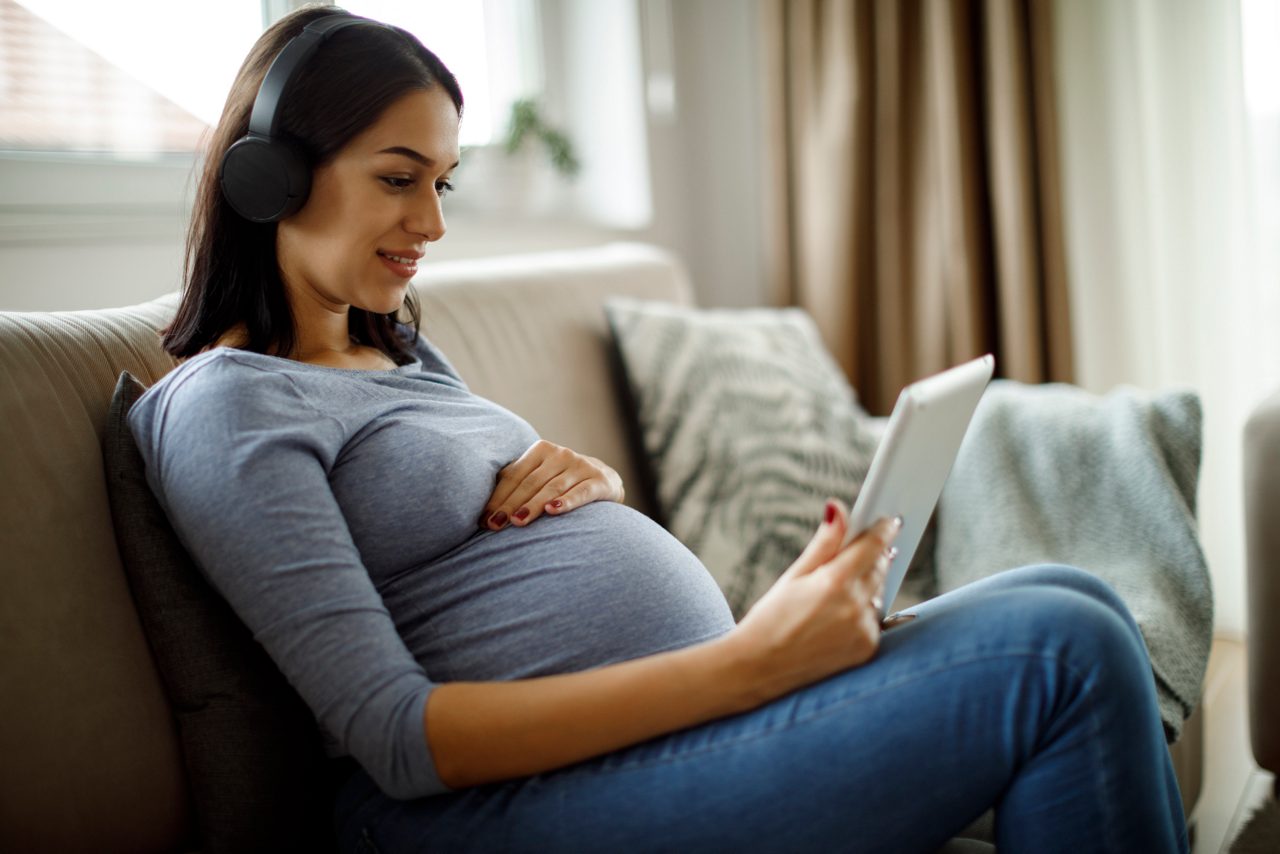 Schwangere sitzt auf der Couch mit Tablet und Kopfhörern