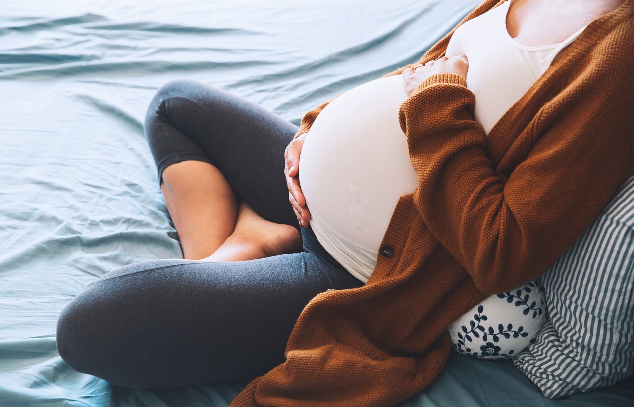 Beautiful pregnant woman sitting at bed and holds hands on belly in bedroom at home. Young mother waiting of a baby. Pregnancy, maternity and expectation image. Close-up, indoors.