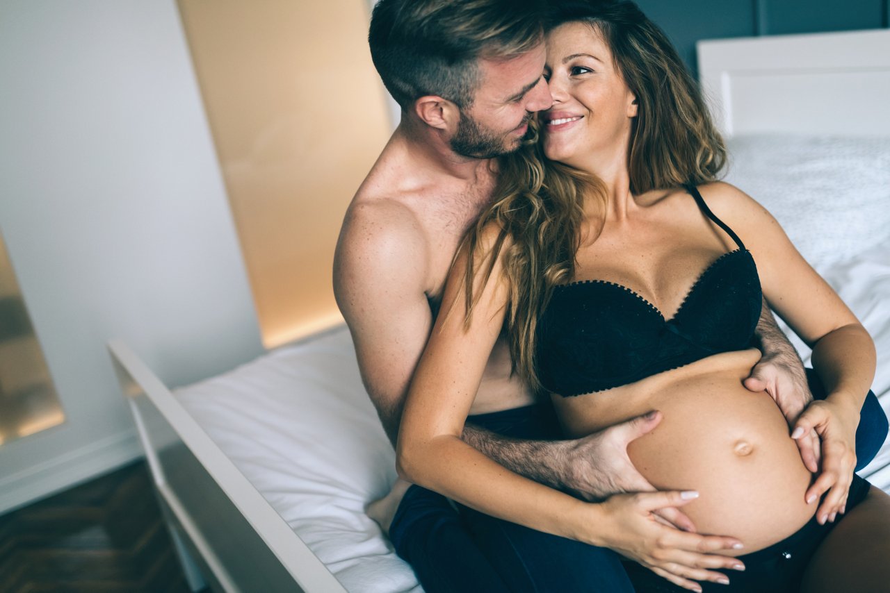 Mann und Schwangere sitzen gemeinsam auf der Couch. Der Mann hält die Frau im Arm und umfasst ihren Bauch.