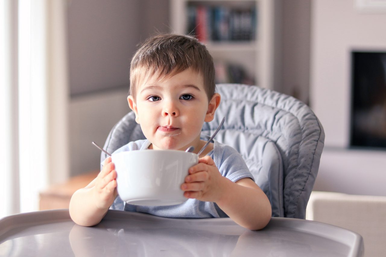 Adorable thankful little baby boy with smeared face just finished his tasty meal and hold white bowl looking at camera wanting more sitting at high feeding chair. Child nutrition concept.