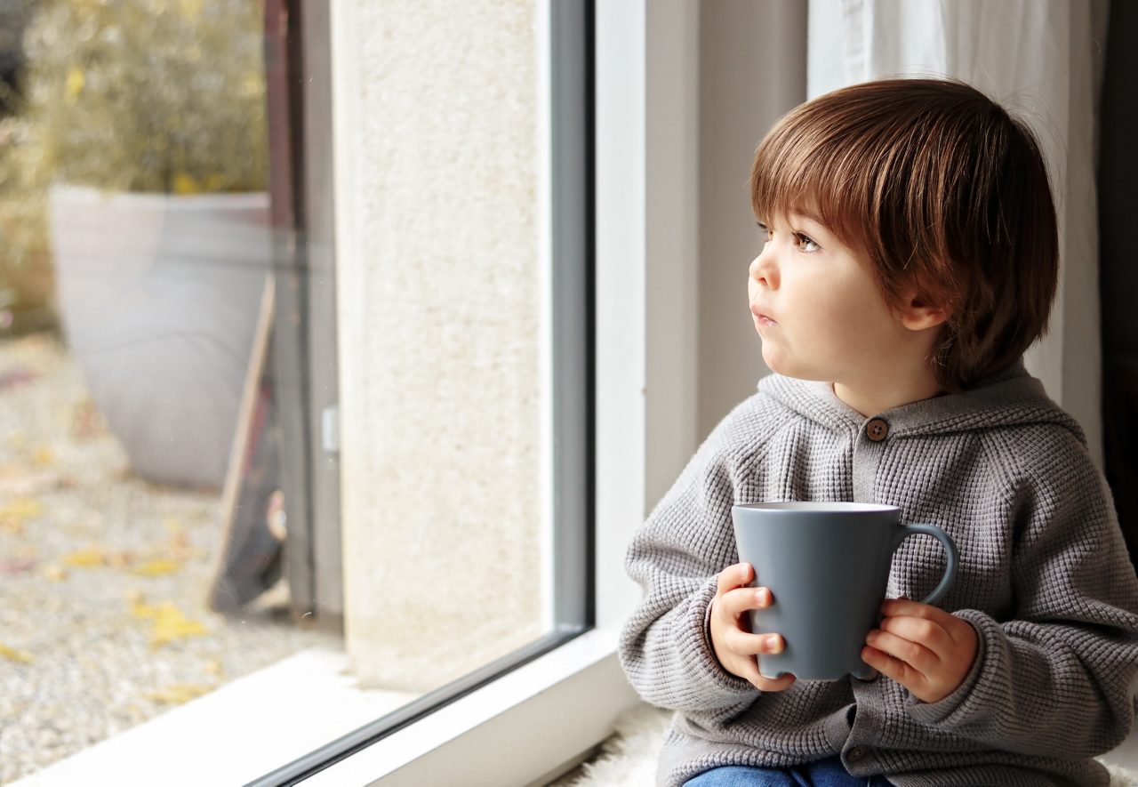 Cute little toddler boy sitting at window with cup of warm tea looking out thoughtfully at chilly autumn weather. Cozy home. Fall melancholy concept. Seasonal mood