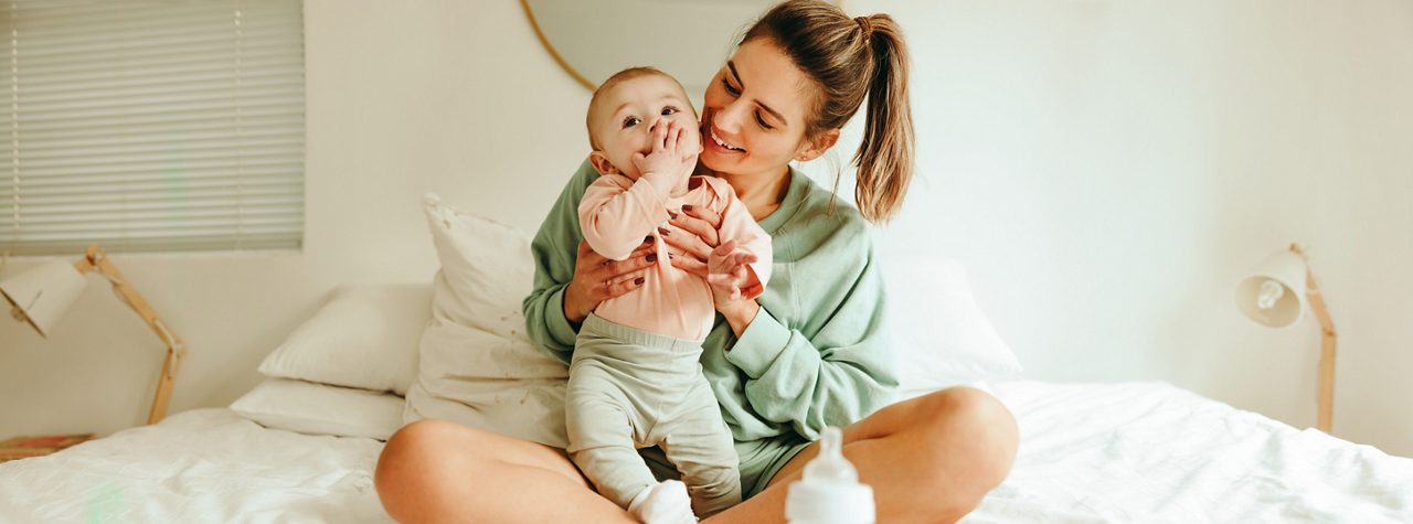 Loving mother smiling at her baby while sitting on the bed at home. Happy young mother caring for her adorable newborn baby. Single mom creating a bond with her infant child.
