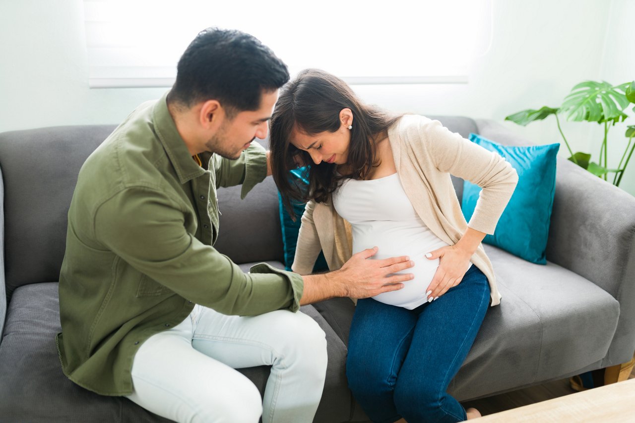 Stressed pregnant woman is having pain in her belly while sitting on the sofa. Alarmed husband trying to help his wife with contractions
