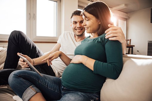 Schwangere Frau und Partner sitzen auf dem Sofa und schauen ein iPad an
