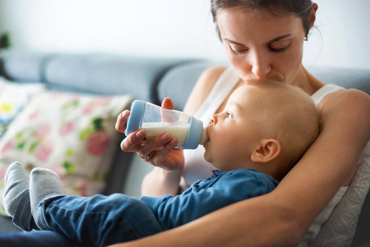 Mutter füttert Baby auf ihrem Schoss und küsst das Köpfchen.