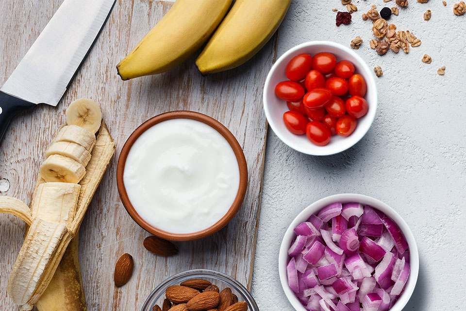Close up image of ingredients on a table: bananas, chopped onion, sour cream, almond, cherry tomatoes