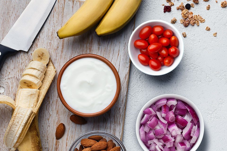 Close up image of ingredients on a table: bananas, chopped onion, sour cream, almond, cherry tomatoes