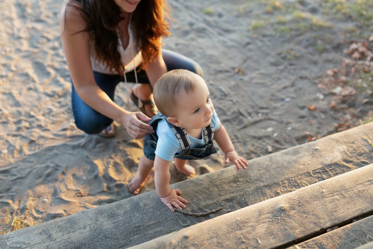 Kleiner Junge hält sich an einer Bank fest. Da hinter hockt die Mama im Sand um zu helfen.