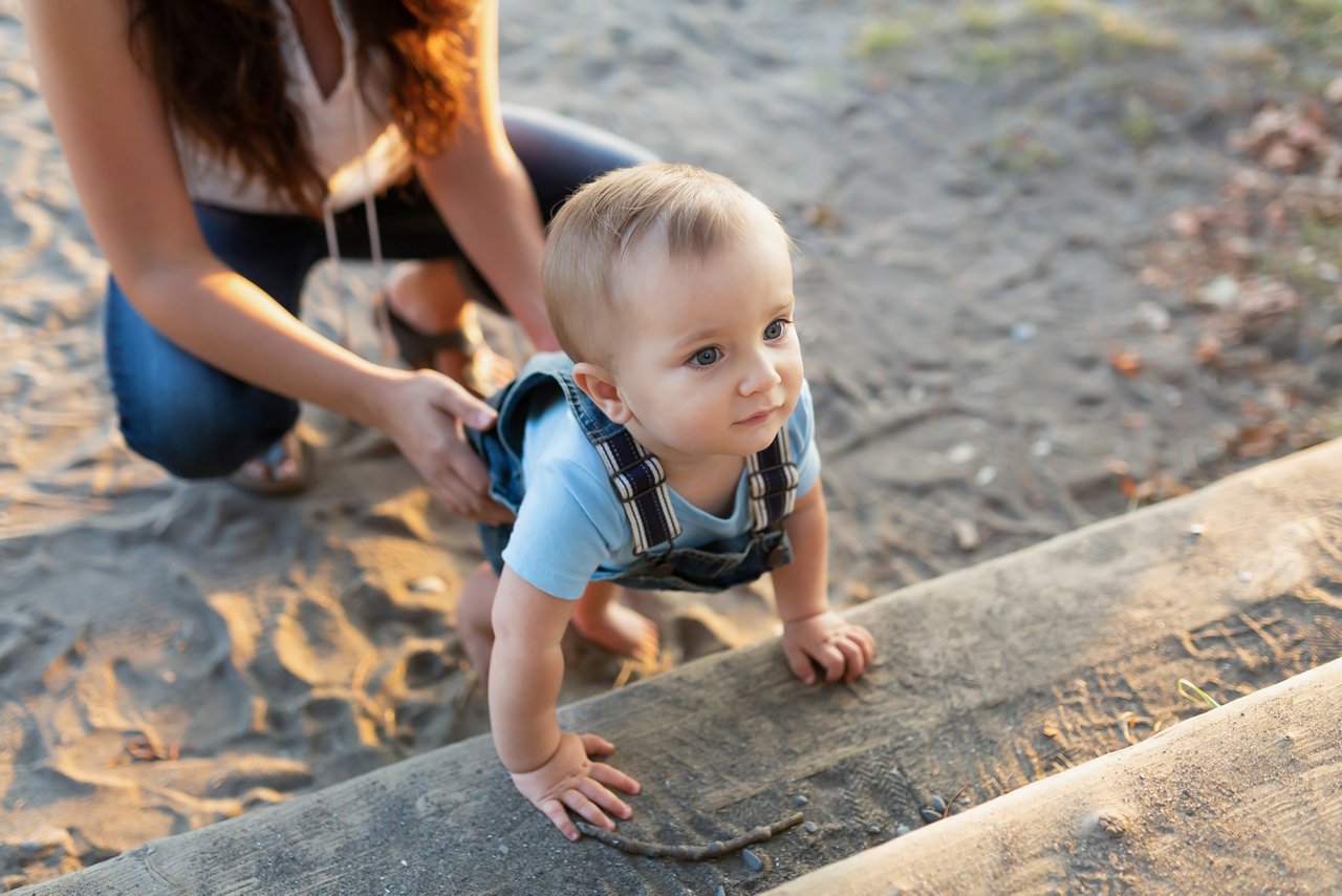 Kleiner Junge hält sich an einer Bank fest. Da hinter hockt die Mama im Sand um zu helfen.