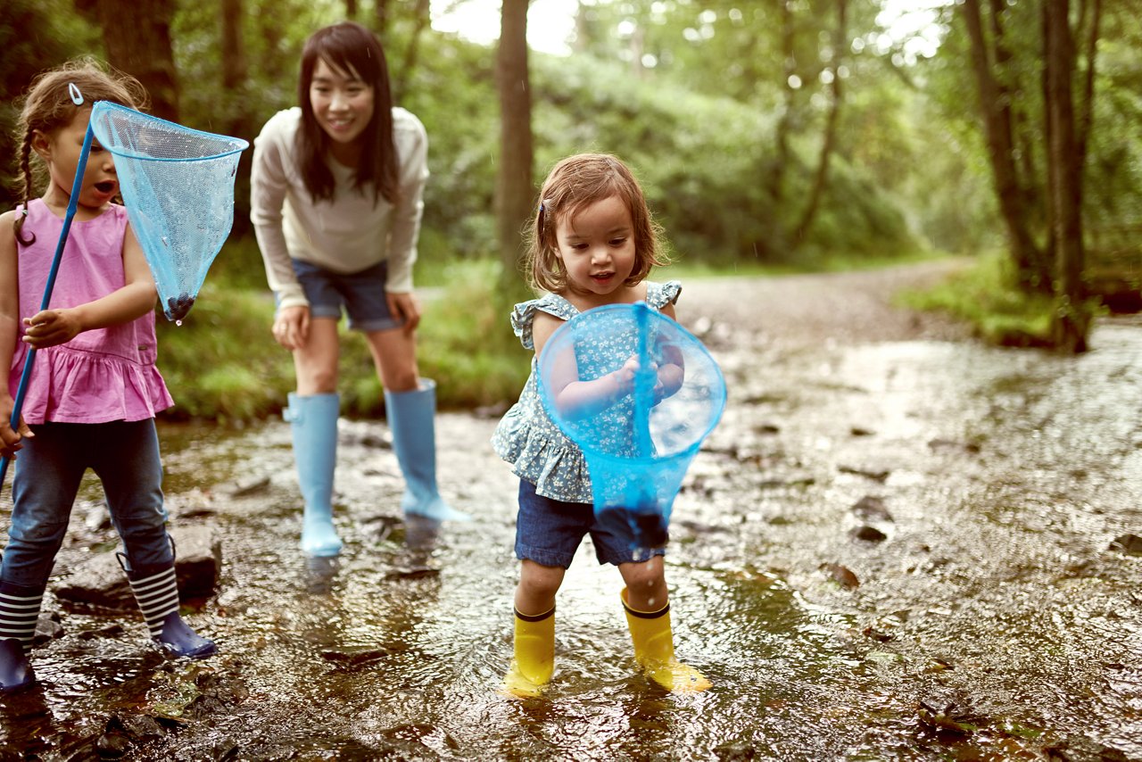 Mama mit zwei Kleindkindern spielt draußen im Matsch. Beide Mädchen haben jeweils einen Kescher in der Hand. Im Hintergrund sieht man einen Wald.