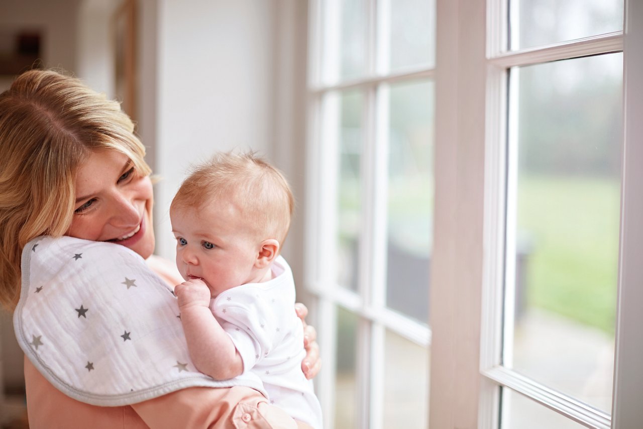 Mama mit Baby auf dem Arm steht am Fenster.