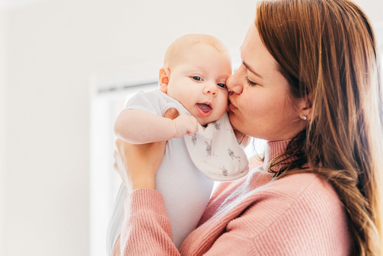 Baby auf Mamas Arm in einer rosa Winterjacke, mit einer weißen Wollmütze auf dem Kopf.