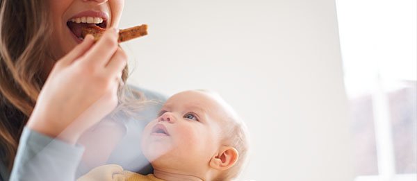 Mutter isst etwas mit Baby auf dem Schoss, das zu ihrem Essen herauf schaut.