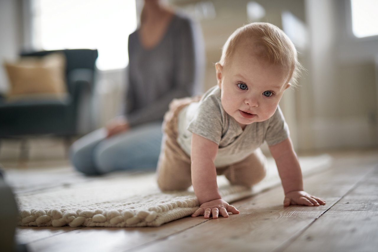 Baby krabbelt durch die Wohnung. Im Hintergrund sitzt eine Frau im Schneidersitz.