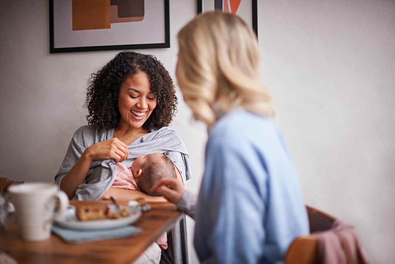 Web only - mum breastfeeding newborn under shirt with friend at cafe table