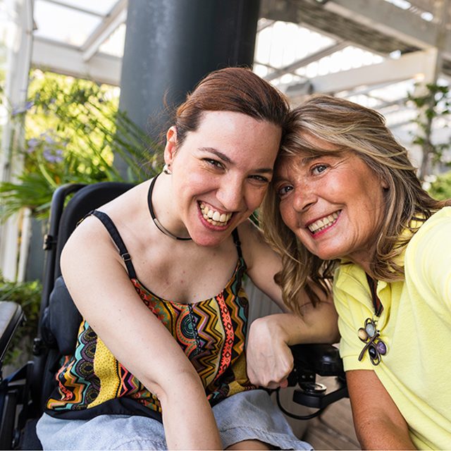 Smiling caregiver and her disabled daughter in a wheelchair are capturing a joyful selfie together, soaking up the warmth of a sunny day outdoors while celebrating their special bond