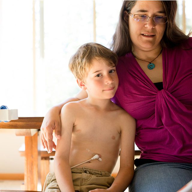 Boy being fed through g-tube smiles at viewer  while mother reads to him