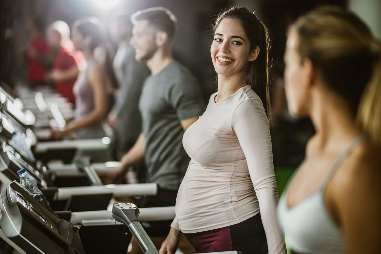 Happy expecting woman exercising on treadmill in a gym and communicating with her friend.