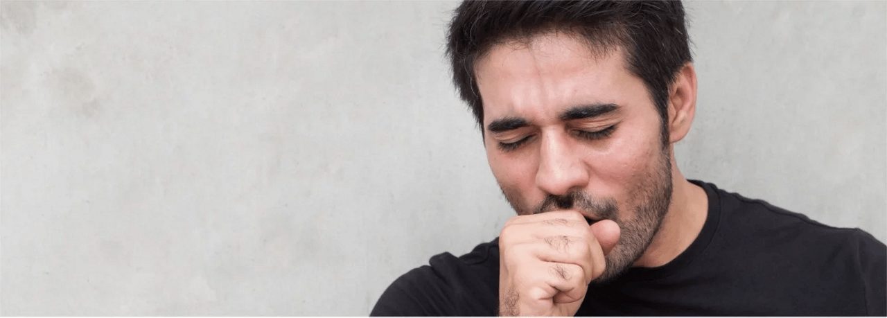 Ein junger Mann hält sich die Faust vor den Mund und hustet, im Hintergrund ist eine graue Wand zu sehen.