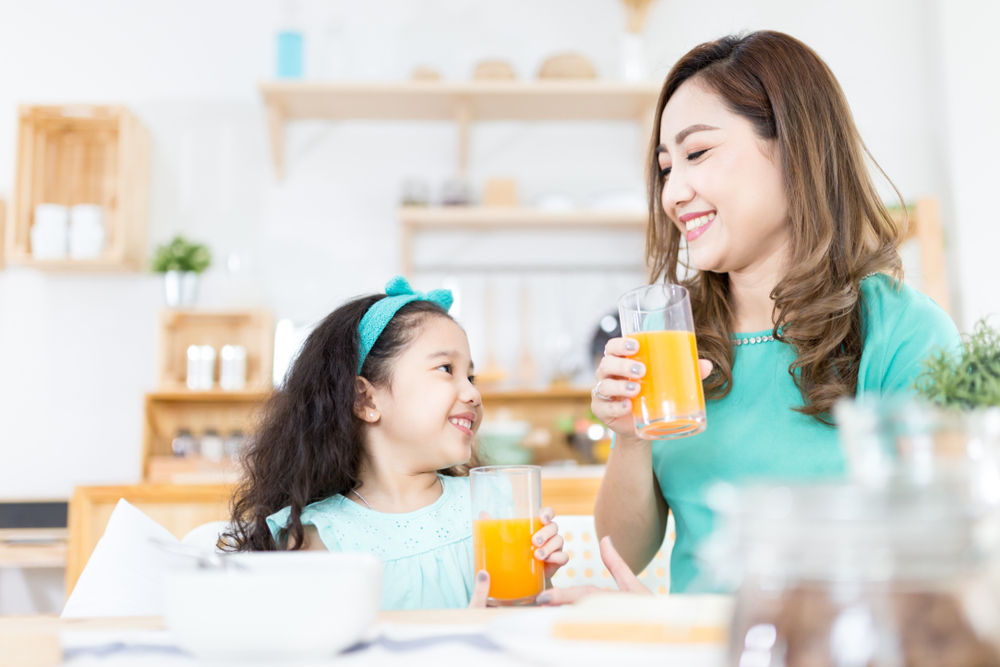 Asian children and her mom drink orange juice in the morning, they holding orange juice glass, child nutrition and development, they feeling happy in family time, health promotion
