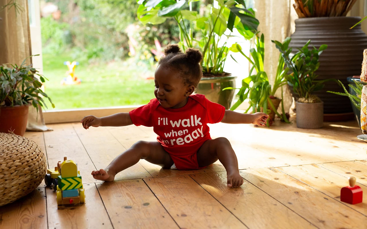 Girl baby playing on a wooden floow wearing a red t-shirt with "I'll walk when I'm ready" printed on it