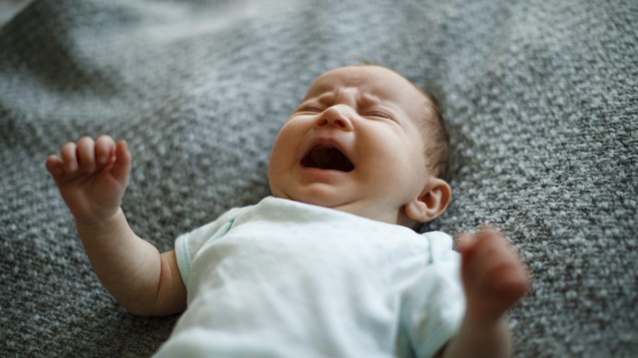 Ein Baby liegt weinend auf einer grauen Decke und trägt einen weißen Strampler.