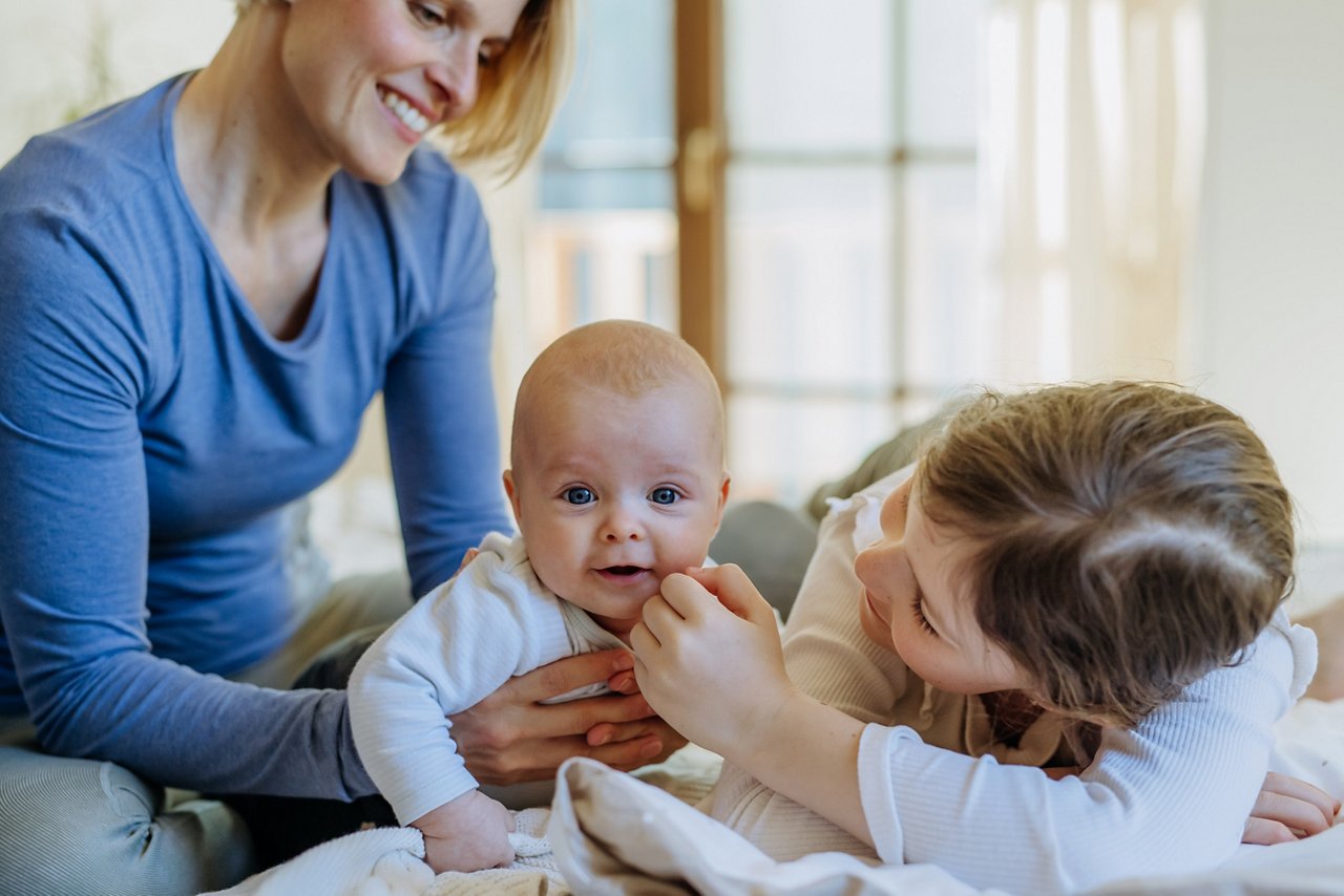 Eine Frau hält ein Baby auf dem Schoß, während ein Kind das Baby streichelt, im Hintergrund ist ein Fenster sichtbar.