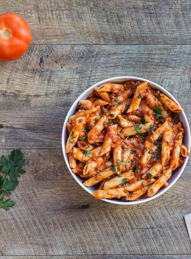 A white bowl filled with penne pasta coated in rich tomato sauce, garnished with fresh parsley. The dish is placed on a rustic wooden surface, accompanied by a ripe tomato and parsley leaves. The vibrant colors of the food create a warm and appetizing visual.