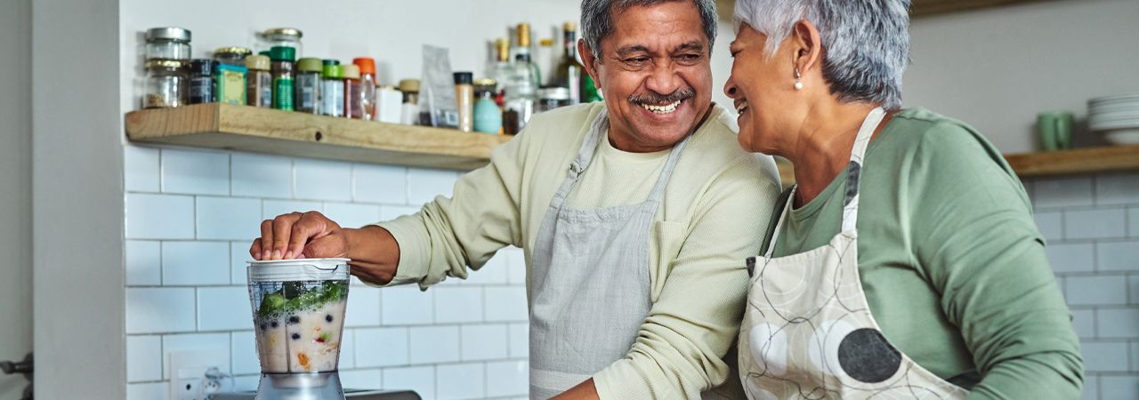 Duas pessoas preparando um smoothie saudável em uma cozinha moderna. O liquidificador está cheio de ingredientes frescos, enquanto vários potes e recipientes de outros ingredientes estão organizados ordenadamente nas prateleiras ao fundo. Esta imagem destaca um estilo de vida vibrante e consciente da saúde.