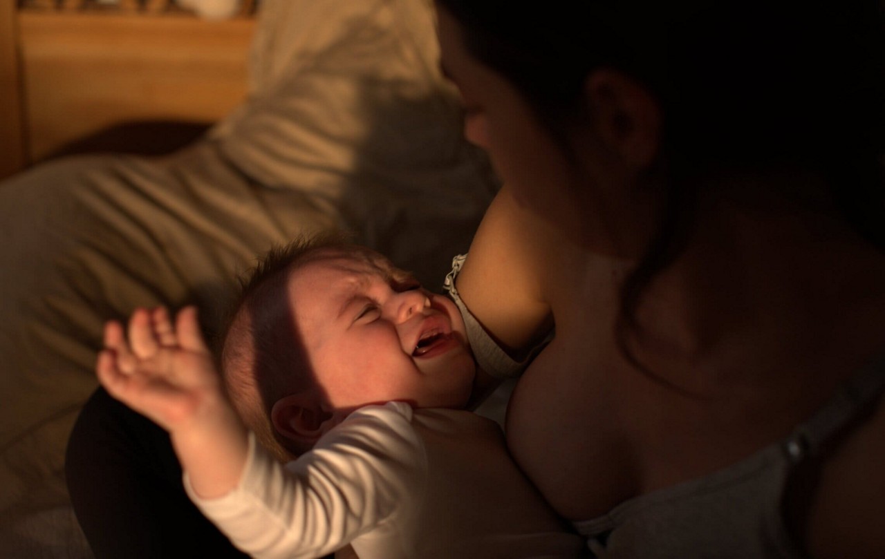 On voit une mere allaiter son bebe dans un cadre interieur confortable. La scene est eclairee par un eclairage doux et chaleureux, creant une atmosphere sereine et intime. Le bebe porte une tenue blanche a manches longues, et l arriere-plan presente un lit avec une literie aux tons neutres.