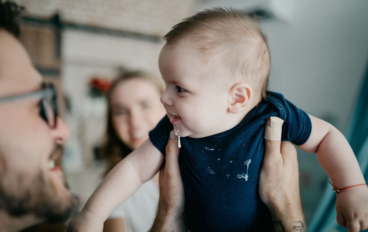 Un bebe portant une grenouillere bleue est tenu par un adulte a l interieur. Le cadre semble etre un environnement domestique confortable avec des elements d arriere-plan flous. Le bebe a les cheveux clairs et porte un petit bandage sur le bras. La grenouillere presente un motif brode subtil.