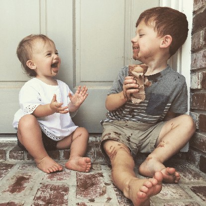 Deux jeunes enfants sont assis sur les marches d un porche en brique dans un cadre exterieur decontracte. L a�ne, vetu d une chemise rayee et d un short, tient une friandise au chocolat partiellement deballee, avec des taches visibles sur ses vetements et sa peau. Le plus jeune enfant, vetu d une tenue blanche, semble faire des gestes avec ses mains. La scene transmet une ambiance ludique et detendue avec des tons chauds et naturels.