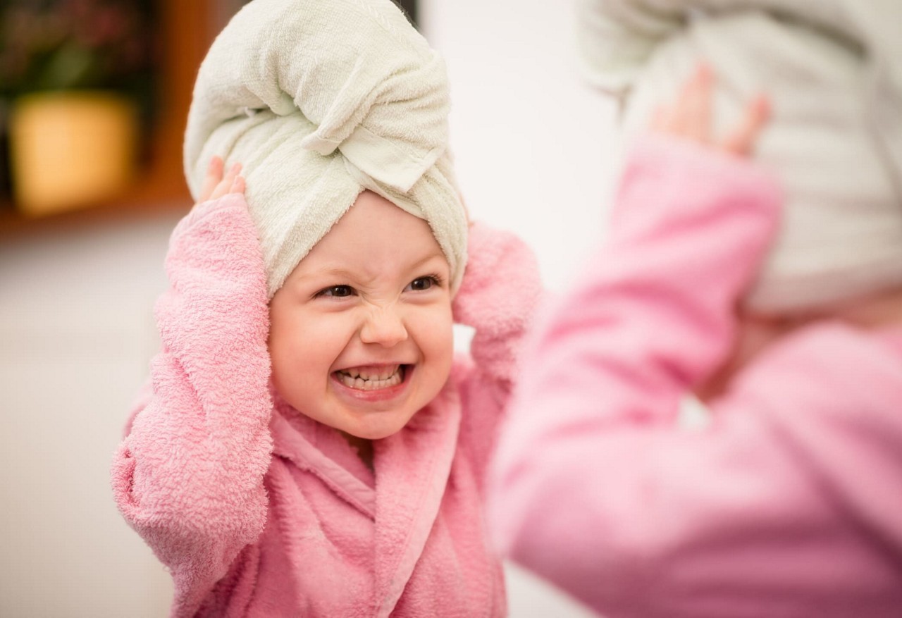 Un jeune enfant vetu d un peignoir rose pale se tient devant un miroir, ajustant une serviette blanche enroulee autour de sa tete. Le decor semble etre a l interieur, probablement une salle de bain, avec des elements d arriere-plan flous. La scene transmet une ambiance chaleureuse et ludique.