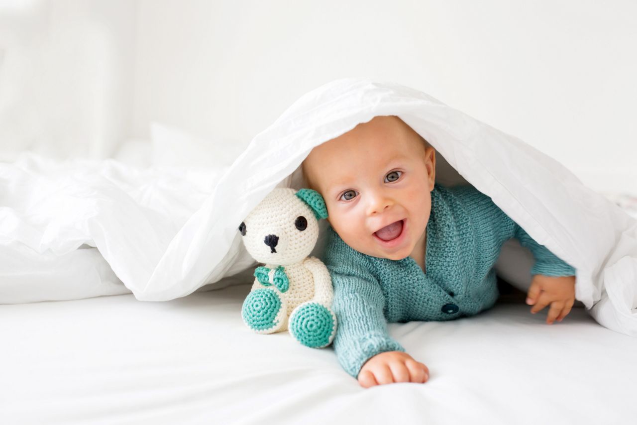 Bebe sorrido embaixo do lencol com ursinho de pelucia