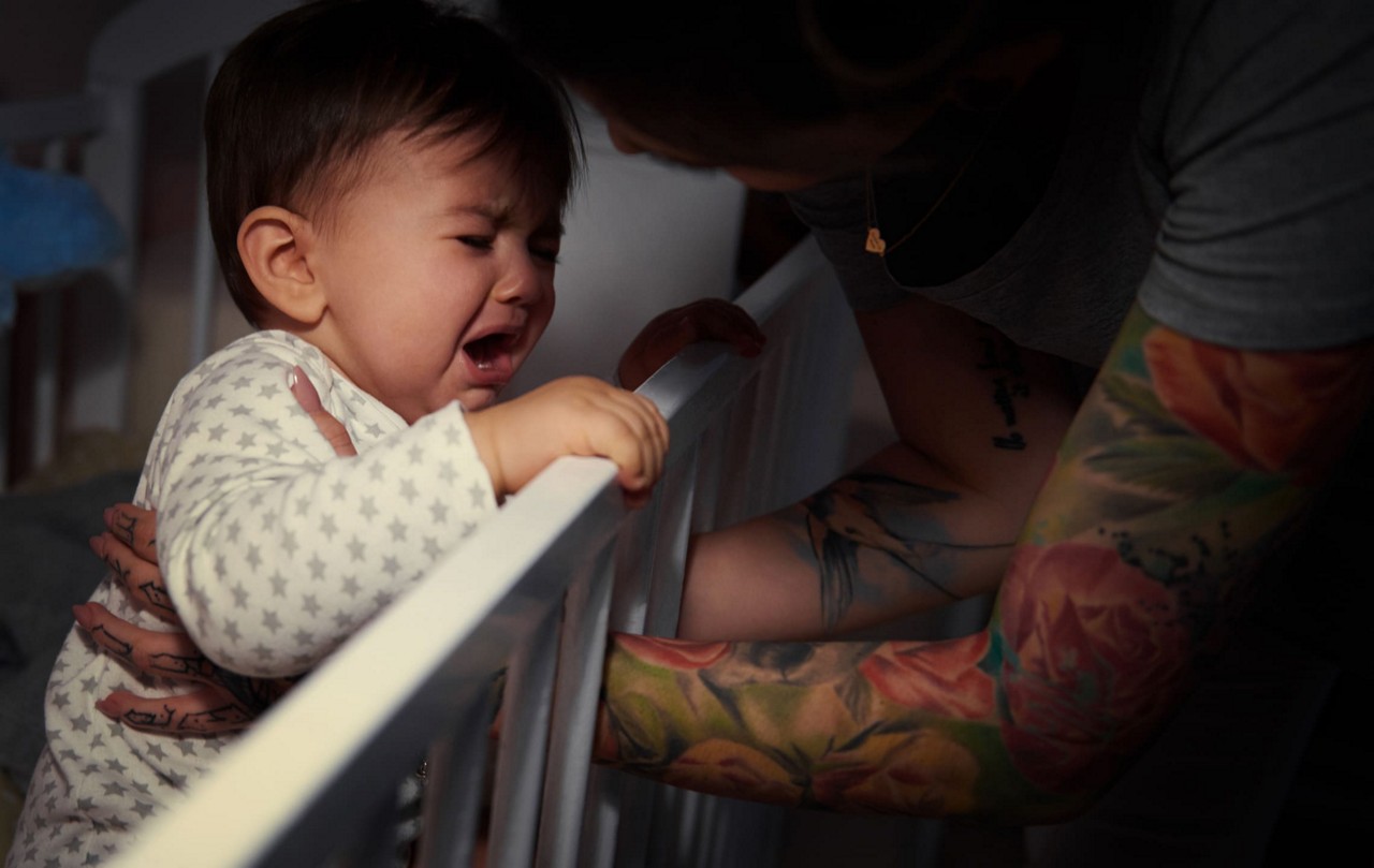 Un adulte tatoue interagit doucement avec un bebe debout dans un berceau. Le cadre semble etre une chambre d enfant doucement eclairee, creant une atmosphere chaleureuse et intime. Le bebe porte une grenouillere a motifs d etoiles et les tatouages floraux colores de la soignante sont bien visibles.