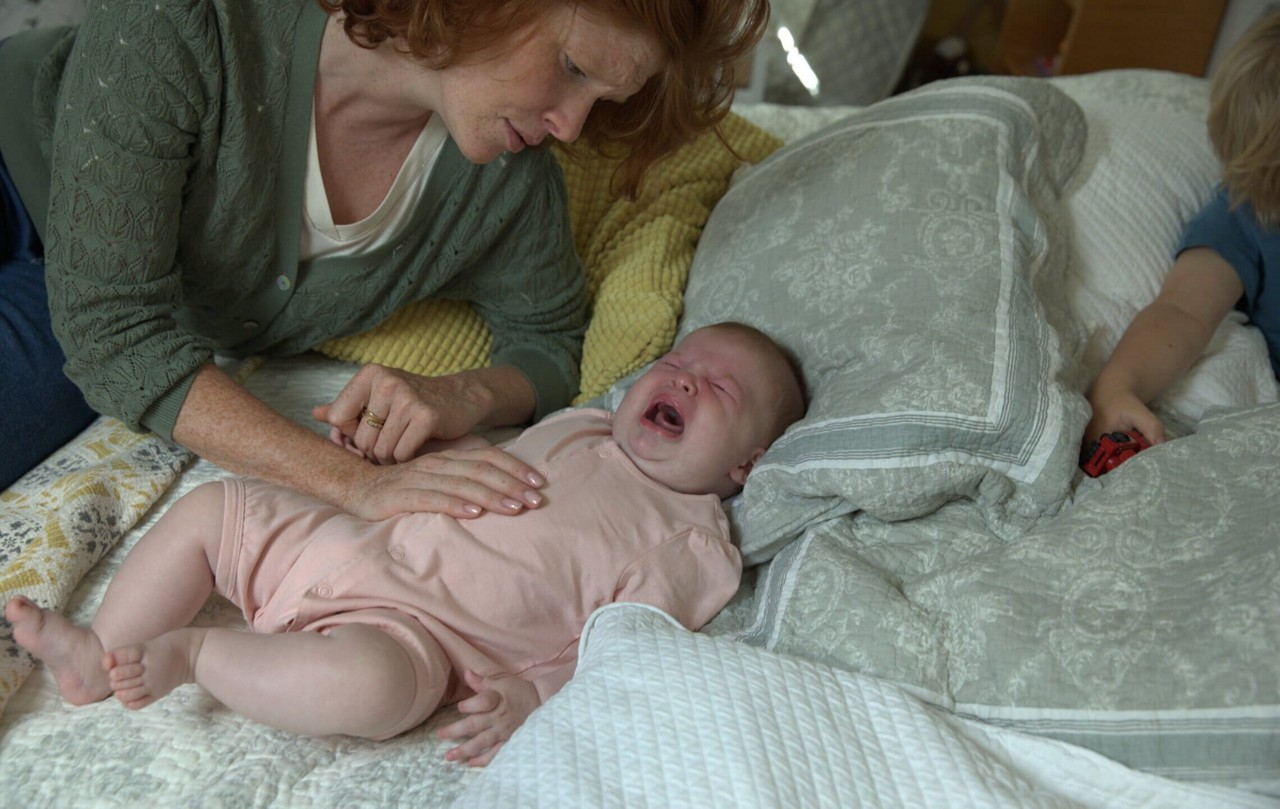 Une femme est assise sur un lit avec un bebe allonge sur une literie moelleuse, vetue d une grenouillere rose clair. Un jeune enfant est visible a proximite, tenant une petite voiture rouge. Le cadre presente des oreillers a motifs et une atmosphere chaleureuse et familiale.