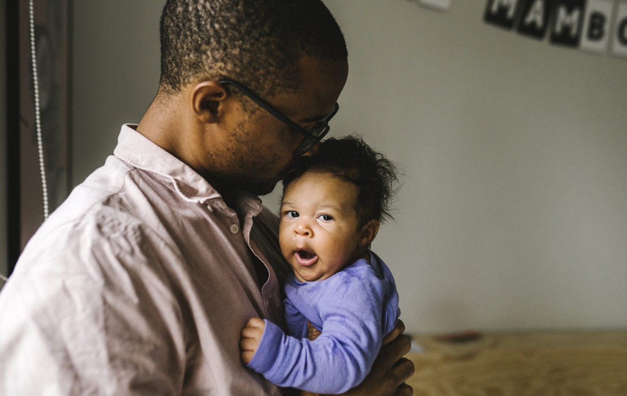 Un homme en chemise rose pale tient un bebe vetu d une grenouillere violette dans un cadre interieur chaud. L arriere-plan presente un eclairage tamise et un decor partiellement visible, y compris une banniere avec le texte � MAMBO �. La scene transmet un moment tendre et intime entre parent et enfant.