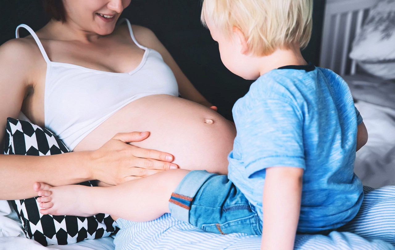 Une femme enceinte est assise sur un lit, tenant doucement son ventre pendant qu un jeune enfant interagit avec elle. Le cadre est intime et chaleureux, avec une literie douce et une atmosphere detendue. Les visuels mettent l accent sur les liens familiaux et les soins maternels, tandis que l eclairage naturel renforce l ambiance chaleureuse.