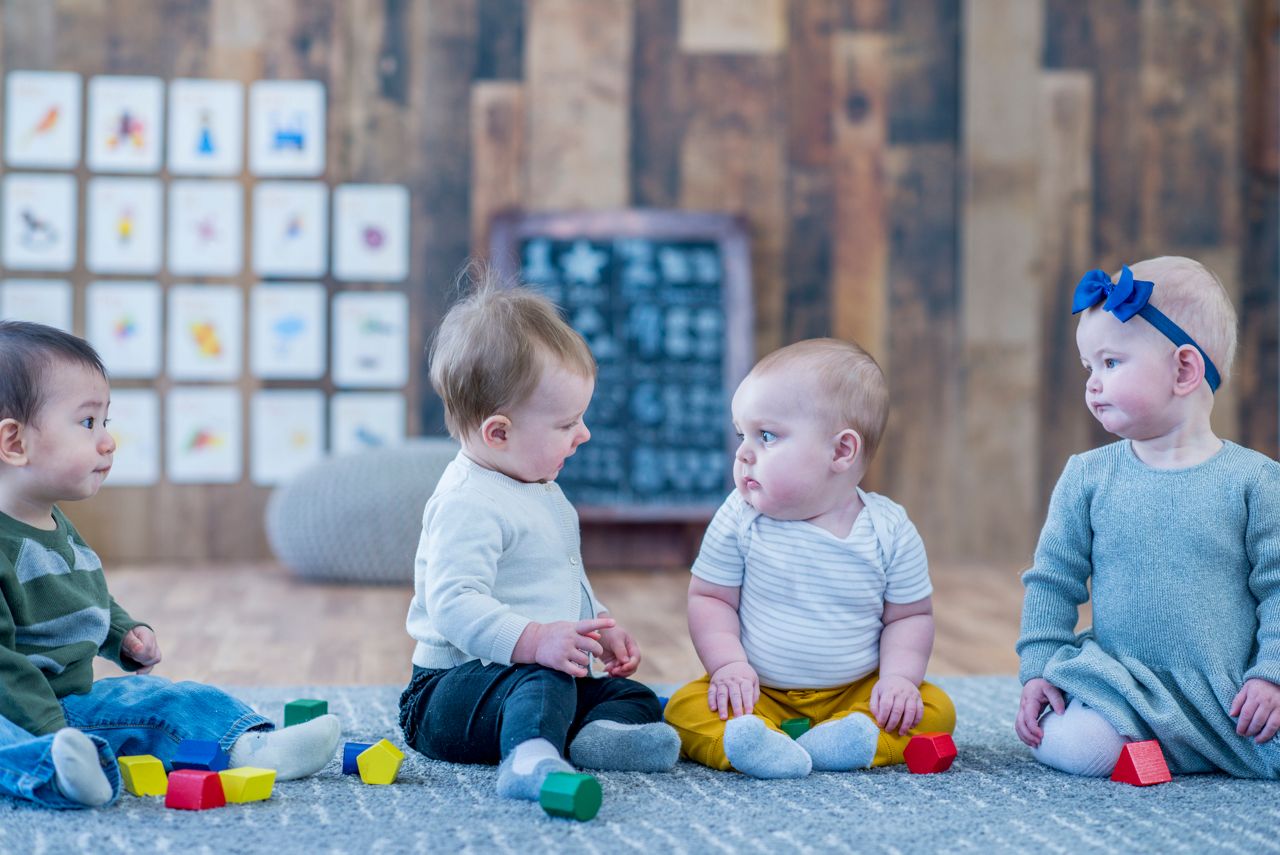 Four babies are indoors in a daycare center. They are all sitting and staring at each other while playing with blocks.