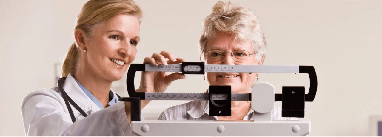 Ärztin misst einer älteren Frau das Gewicht mit einer mechanischen Waage in einer Praxis, beide lächeln.