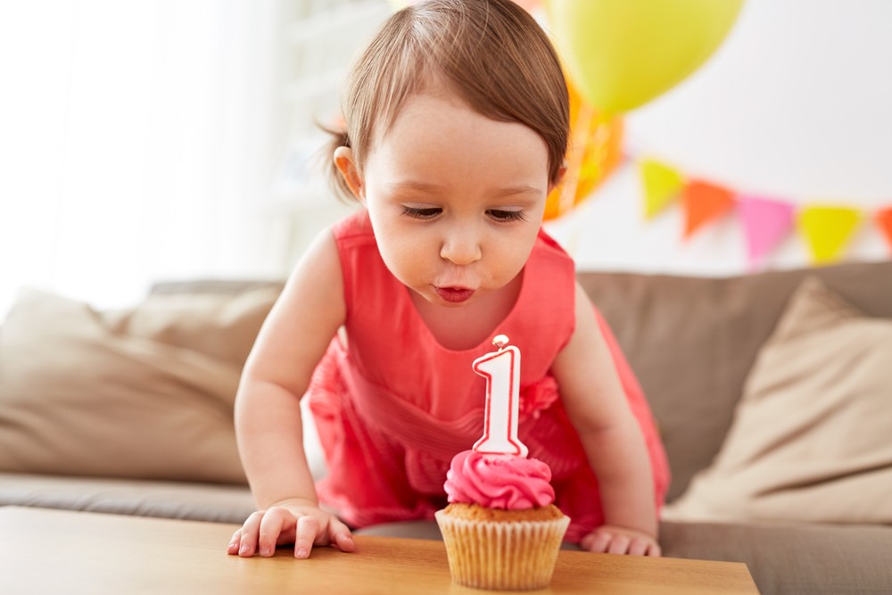 childhood, people and celebration concept - happy baby girl blowing to candle with number 1 on cupcake on birthday party at home