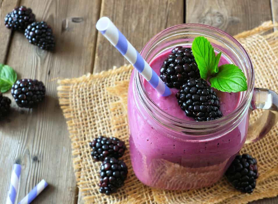 A vibrant purple blackberry smoothie served in a mason jar with a striped straw. Fresh blackberries and mint leaves garnish the drink, placed on a rustic burlap mat over a wooden table. The setting evokes a natural and wholesome vibe, perfect for healthy living.