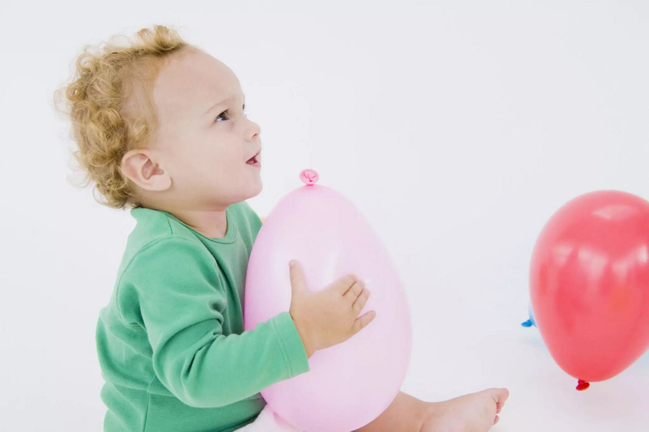 Un jeune enfant vetu d'une tenue verte est assis a l'interieur, tenant un ballon rose. Un autre ballon rouge est visible a proximite sur le sol blanc. Le decor est minimaliste et lumineux, mettant l'accent sur des images ludiques et joyeuses.
